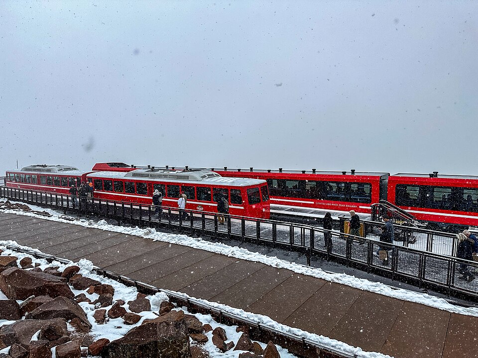 Red cog trains at the snowy Pikes Peak summit platform, with passengers on the walkway.
